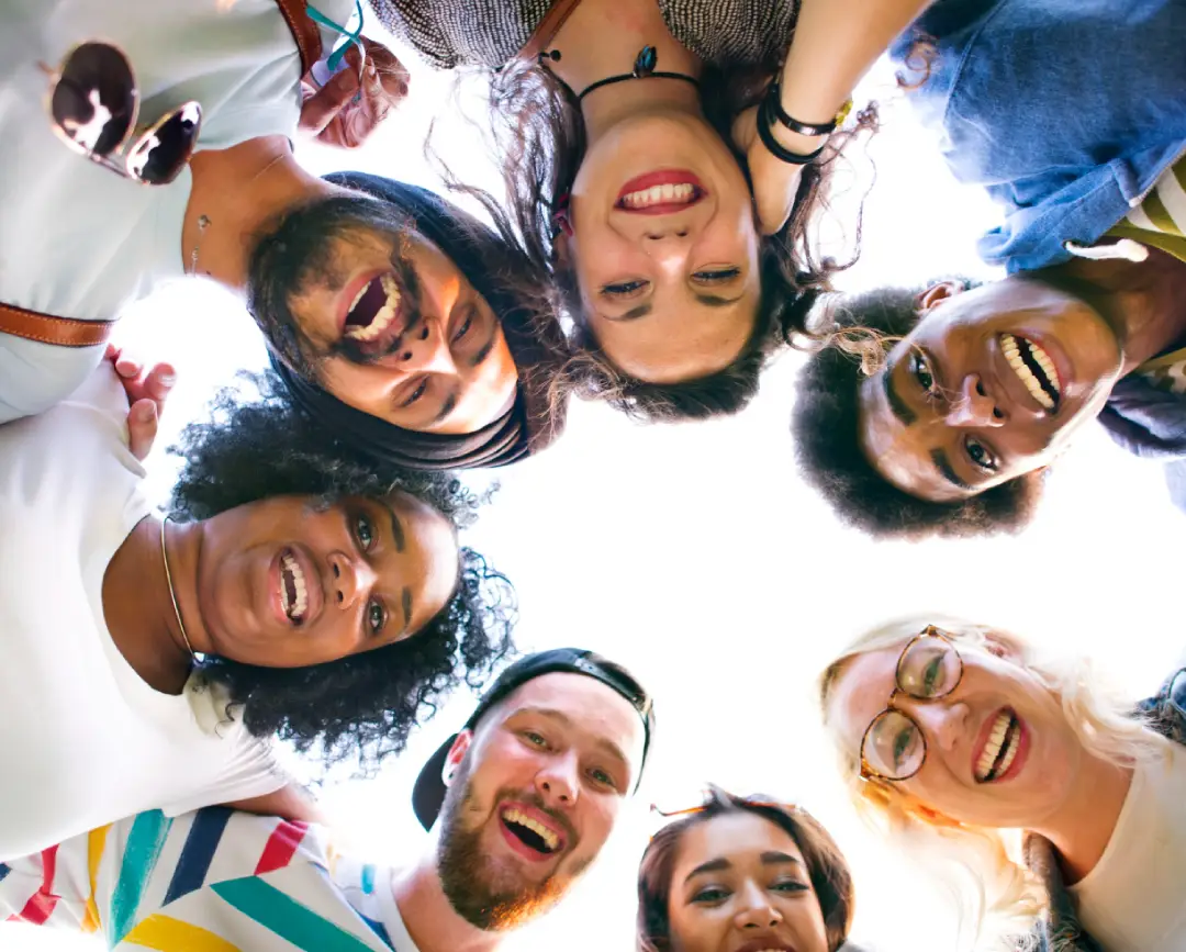 A group of diverse individuals gathers in a circle, looking down with smiles, illuminated by natural light from above A group of diverse individuals gathers in a circle, looking down with smiles, illuminated by natural light from above