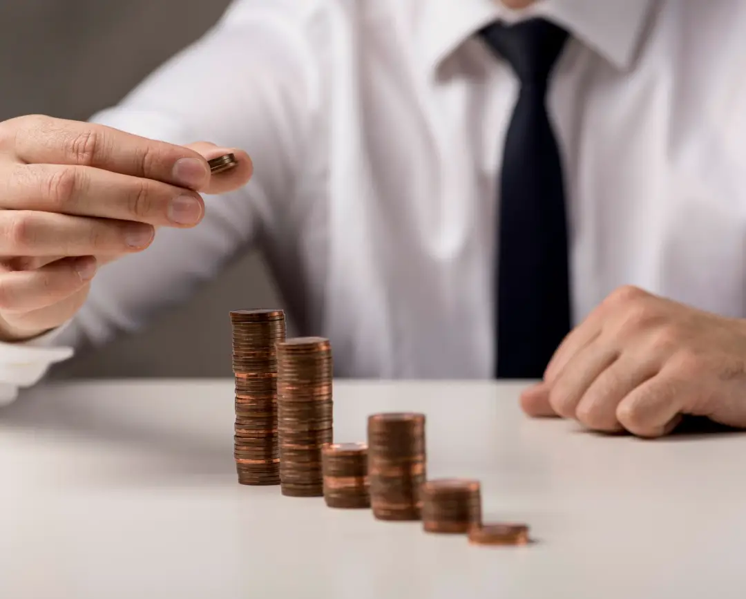 A man in a white shirt and tie stacks coins, illustrating financial growth, with several ascending towers of pennies in front of him A man in a white shirt and tie stacks coins, illustrating financial growth, with several ascending towers of pennies in front of him