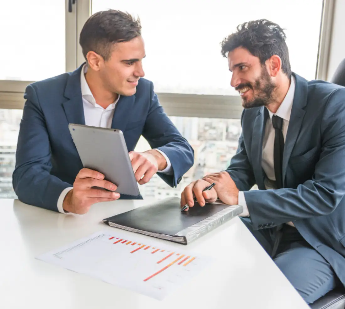 Two business professionals in formal attire are engaged in a discussion at a modern office table with a city skyline view, one using a tablet Two business professionals in formal attire are engaged in a discussion at a modern office table with a city skyline view, one using a tablet